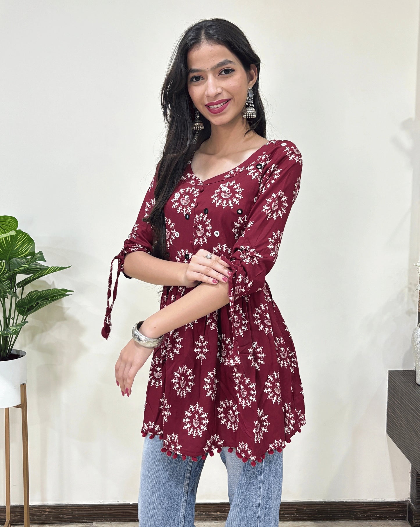 Close-up of Red Woman's Ajrakh Vegetable dyed plant based cotton Short Kurti with beautiful geometric Warli print, frock style pattern, Round neck with Hand-mirror work, lace on sleeves, paired with light blue jeans, green leaf post on right side & Standing in front of white wall.