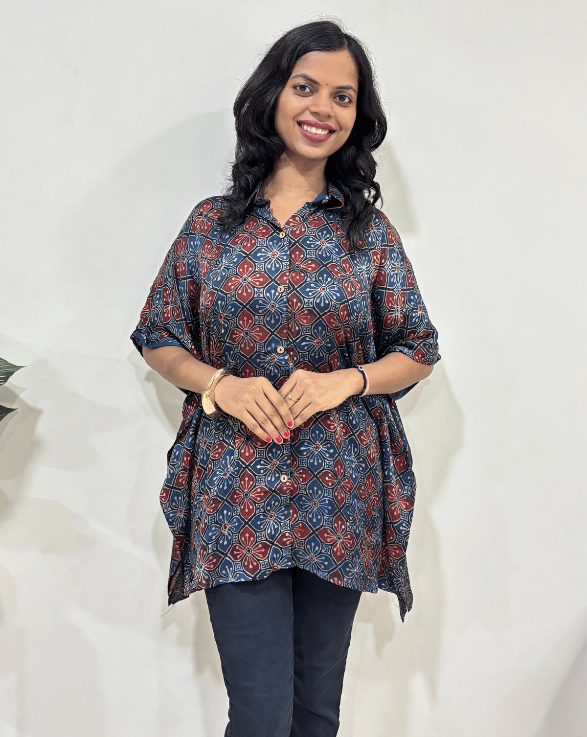 Front view of Deep Blue Women's Ajrakh pure Mashru Silk Short Kaftan Shirt Featured with Collar neck & Elbow sleeves with a bold abstract of White, black, and red geometric circles print, paired with dark pants, standing in front of a white wall.
