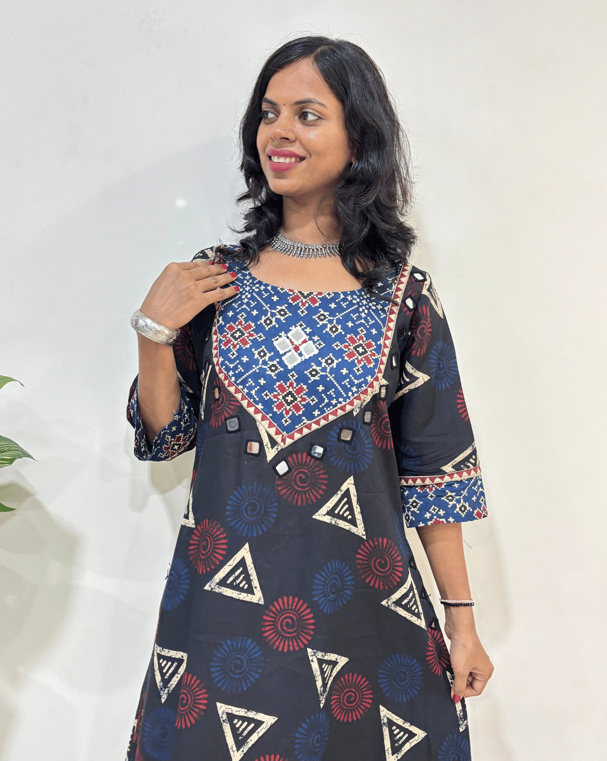 Close-up of Black Women's Ajrakh Vegetable dyed plant based cotton One-peice Featured three-quarter Sleeves & Round Neck, with beautiful patola print yoke on front, bottom & Sleeves , Geometric print on front & sleeves Hand-mirror work on front chest, pocket & Sleeves cuff, Standing in front of White wall.