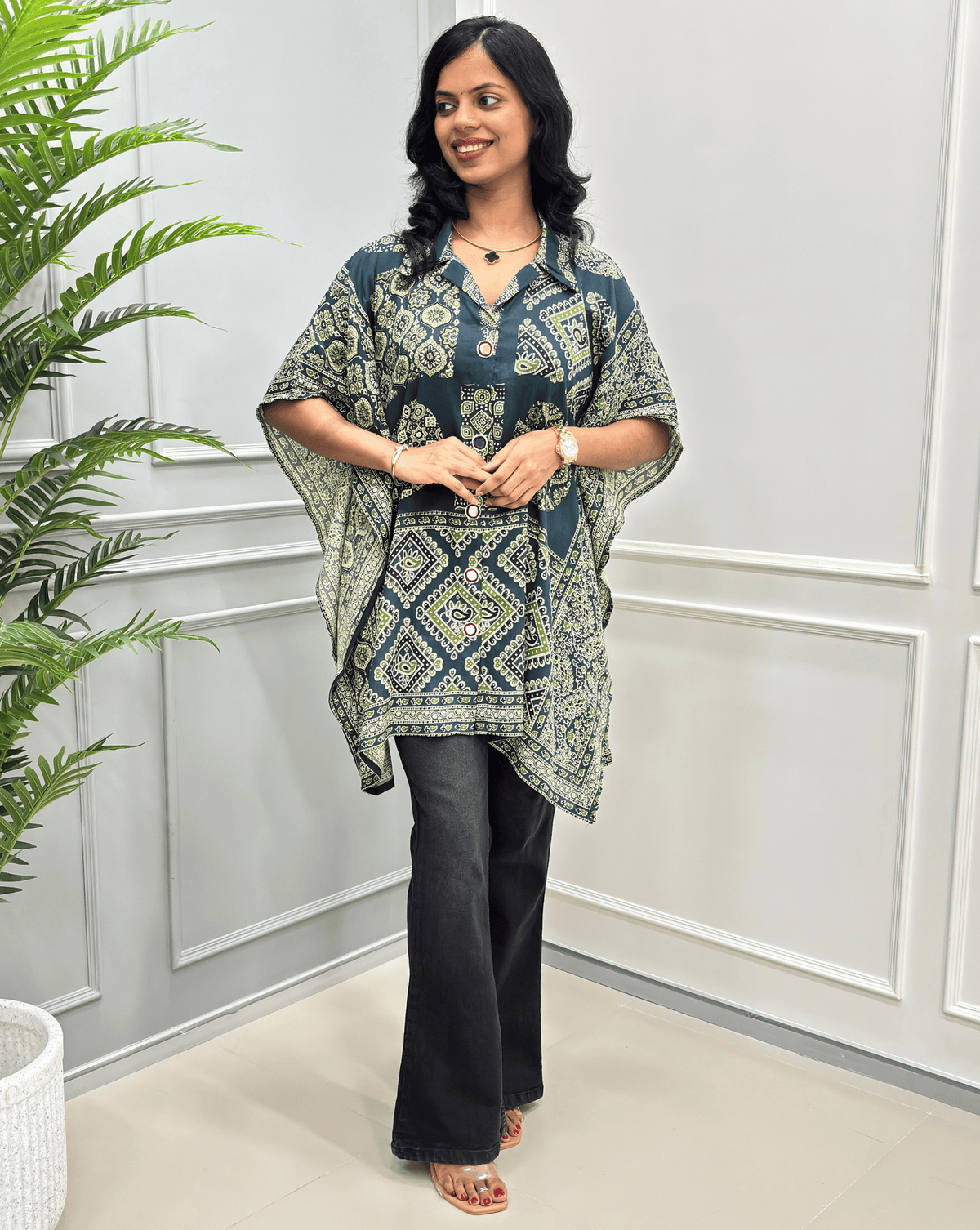 A printed tunic top with wide sleeves, paired with black flared pants, standing in a bright indoor setting with a white paneled wall and a green plant on the left.