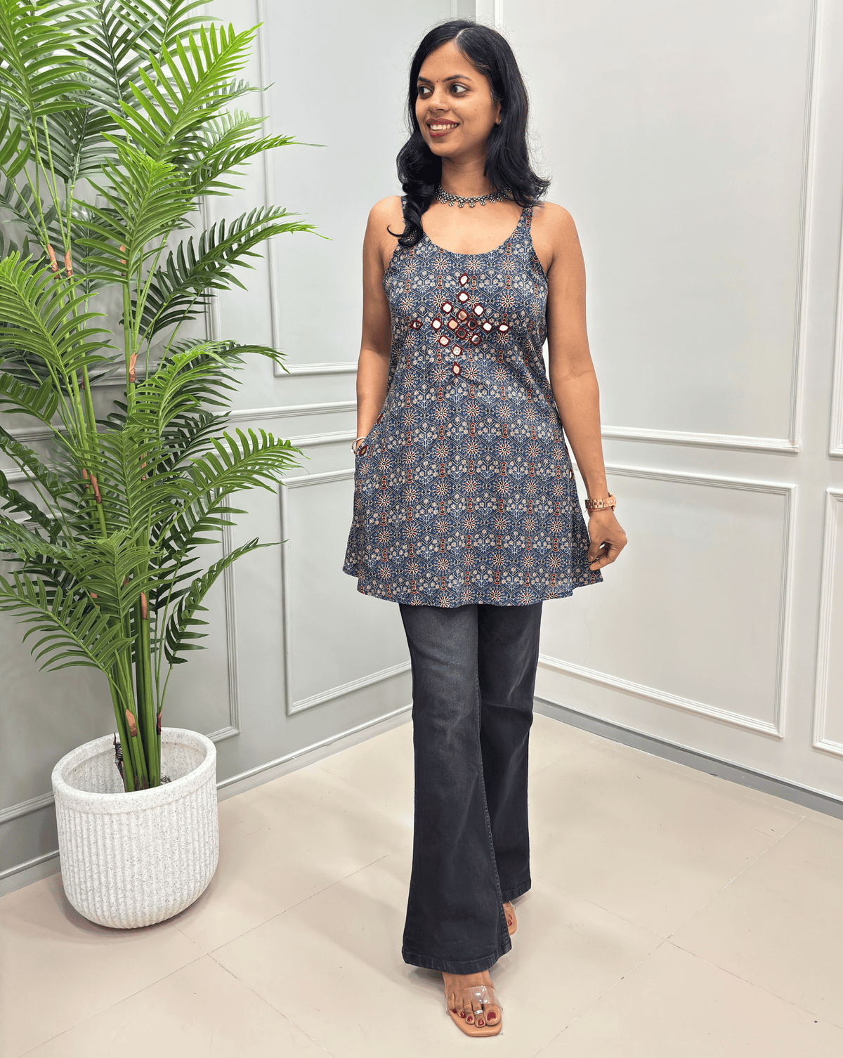 Blue women's Ajrakh cotton sleeveless, Short tunic top front view with a blue and red ajrakh floral pattern, deep round neck, hand-mirror work on chest paired with dark grey flare pants.