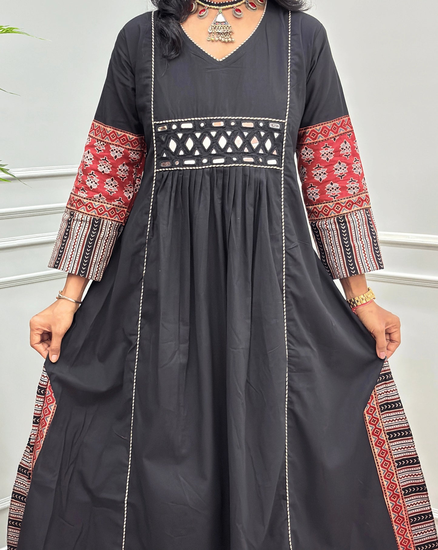 Close-up of handcrafted black Ajrakh cotton long kurti with red and beige patterned sleeve cuffs, a matching patterned waistband, and white embroidered vertical lines, shown against a light grey indoor wall.