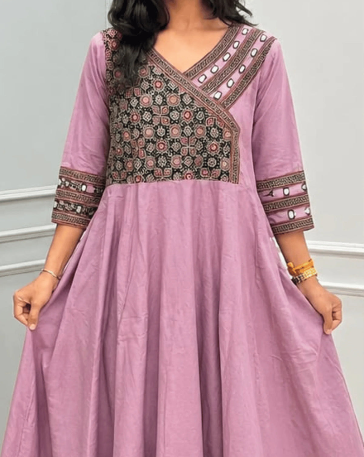 Close-up Veiw of Onion Pink Women's Ajrakh Pure Cotton A-line Anarkali Featured with V-neck with a Black base & Red floral yoke patterned along Real Hand-mirror work on Front & sleeve cuffs , 2-Side pockets, standing on a round woven rug in front of a light gray paneled wall.
