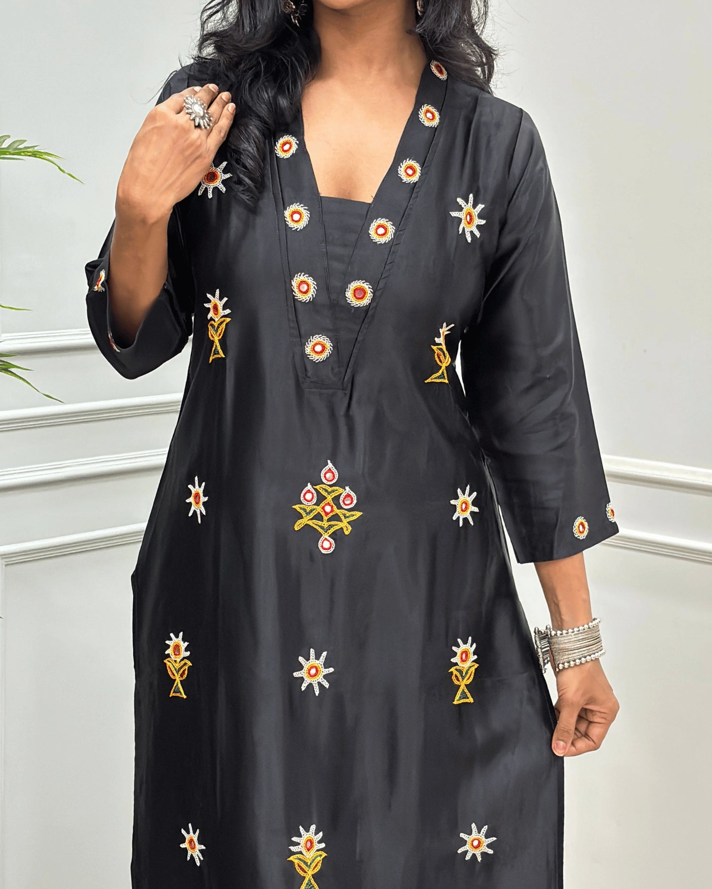 Close-up view of Black Mashru Silk Long Kurti with colorful embroidered floral motifs and a Hand-mirror work on V-neckline, Front & Cuffs, Full Sleeves , standing against a light gray paneled wall.