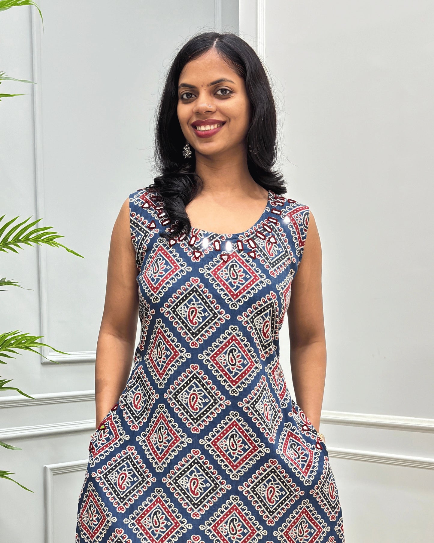 Close-up of Blue Women's cotton long sleeveless Kurti with a red, white, and beige geometric diamond pattern, Hand-mirror work on rounnd neck line border, standing against a white paneled wall.