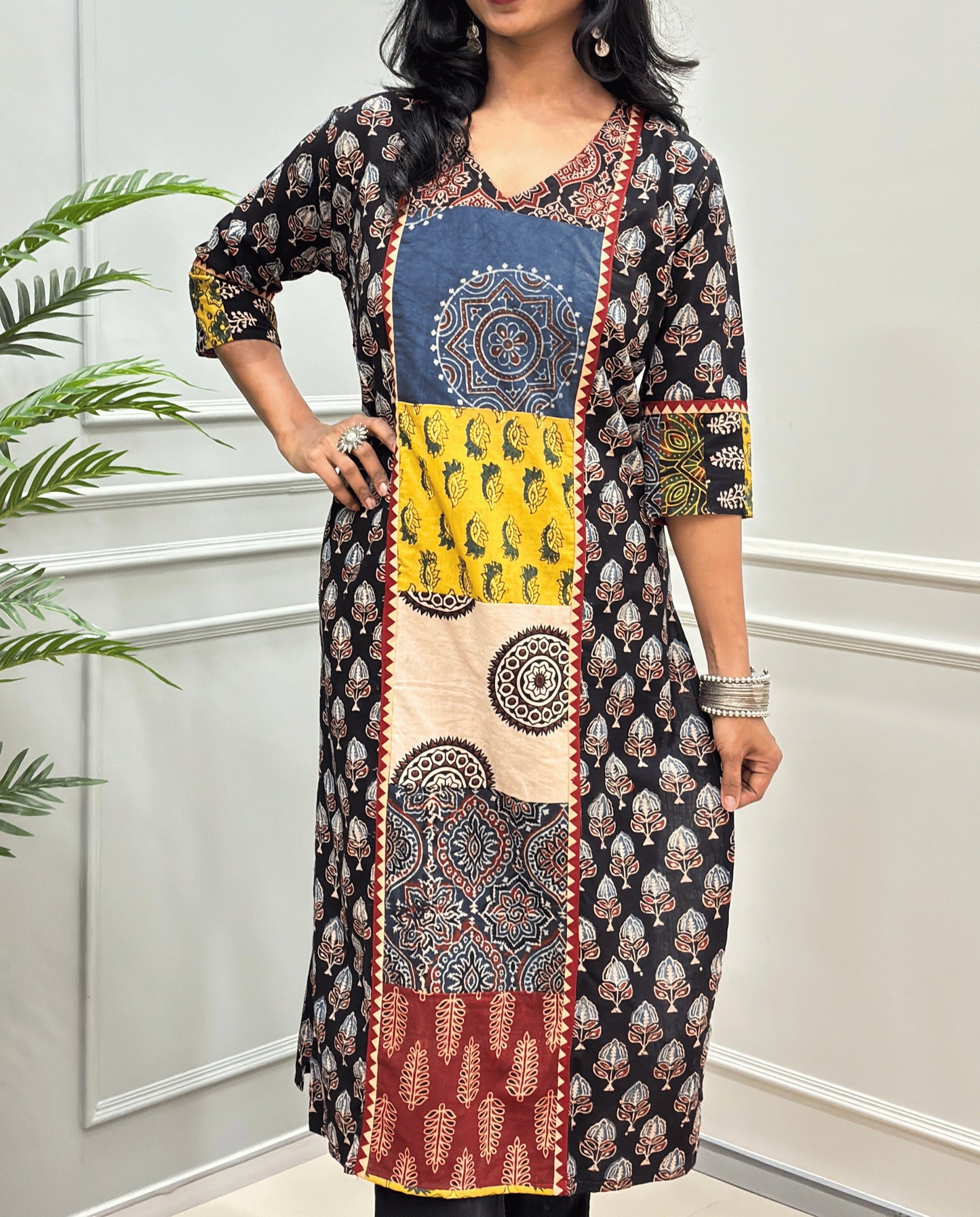 Close-up of Black Women pure cotton long kurti featured with blue, yellow, red, and black Geometric different print panels on Front & Sleeves cuffs , Ajrakh print on blue base outside the middle panel , three quarter Sleeves & V-neck standing indoors against a light paneled wall with a green plant on the left.