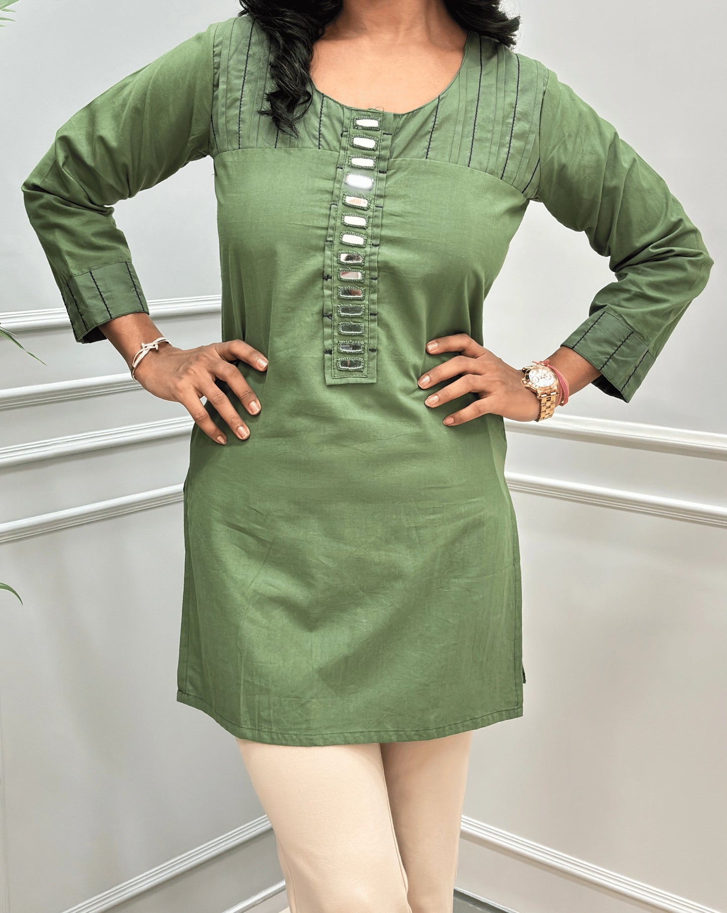 Close-up of Teal-Green women's Ajrakh Vegetable Dyed plant based Cotton Short-kurti with Beige piping and Hand-mirror work look a like button details, Round neck & Full Sleeves, paired with beige Flared pants, standing in a room with a light gray paneled wall and a green plant on the left.