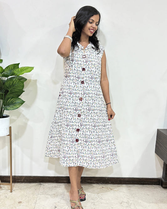 White Women's Ajrakh A-line Vegetable Dyed plant based cotton One-peice with a multi colour Warli print , standing against a white wall with a plant to her left.