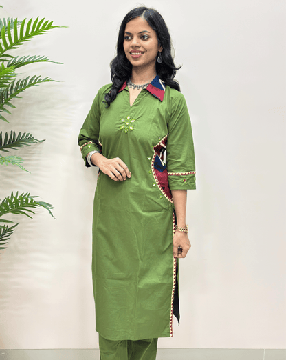 front view of ajrakh long kurti with multicolor geometric patterned accents on the collar, cuffs, and side panel, paired with matching green trousers, set against a plain light wall with a decorative plant on the left.