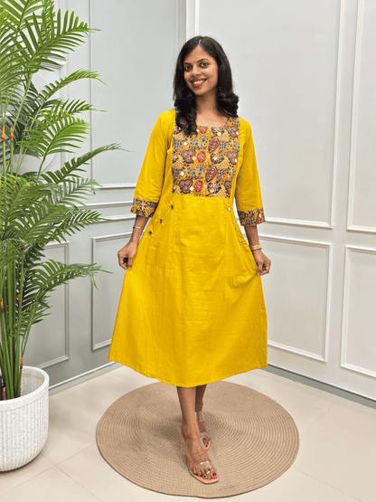  Woman wearing a bright yellow A-line kurta with a colorful embroidered yoke and patterned sleeve , standing on a round woven mat in front of a grey-white paneled wall with a potted plant.