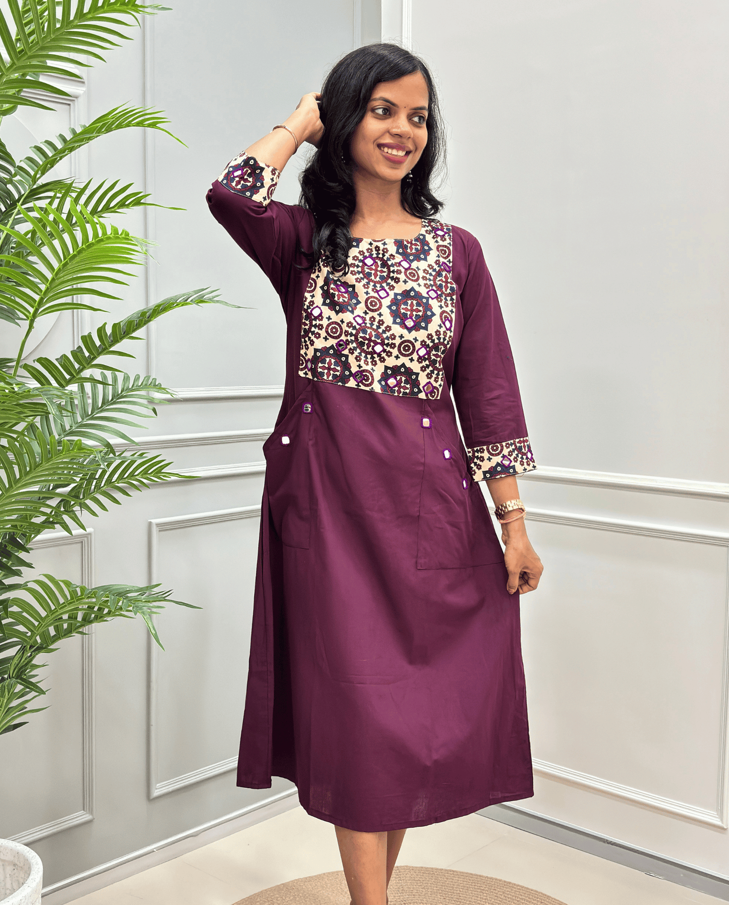  a Women's three-quarter sleeve one-peice dress in deep purple, featuring a colorful geometric pattern and matching patterned cuffs on end of the sleeves, standing in a room with a white paneled wall and a green plant on the left.