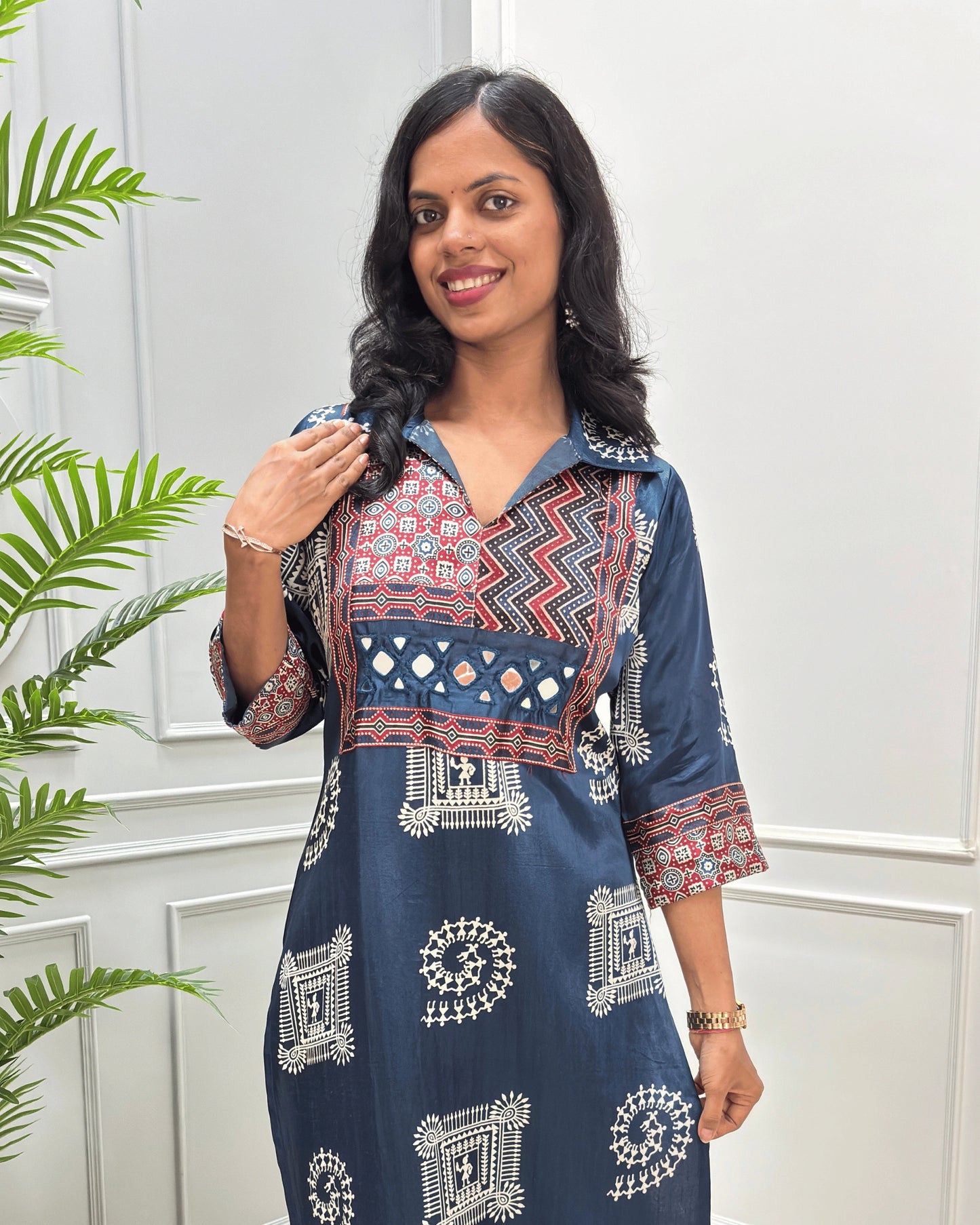 Close-up of Navy Blue Ajrakh Mashru silk Long kurti with multicolor geometric , warli print on front & sleeves along eith hand -mirror work on front , Collar V shaped neck patterns, standing in front of a white paneled wall with a green plant on the left.