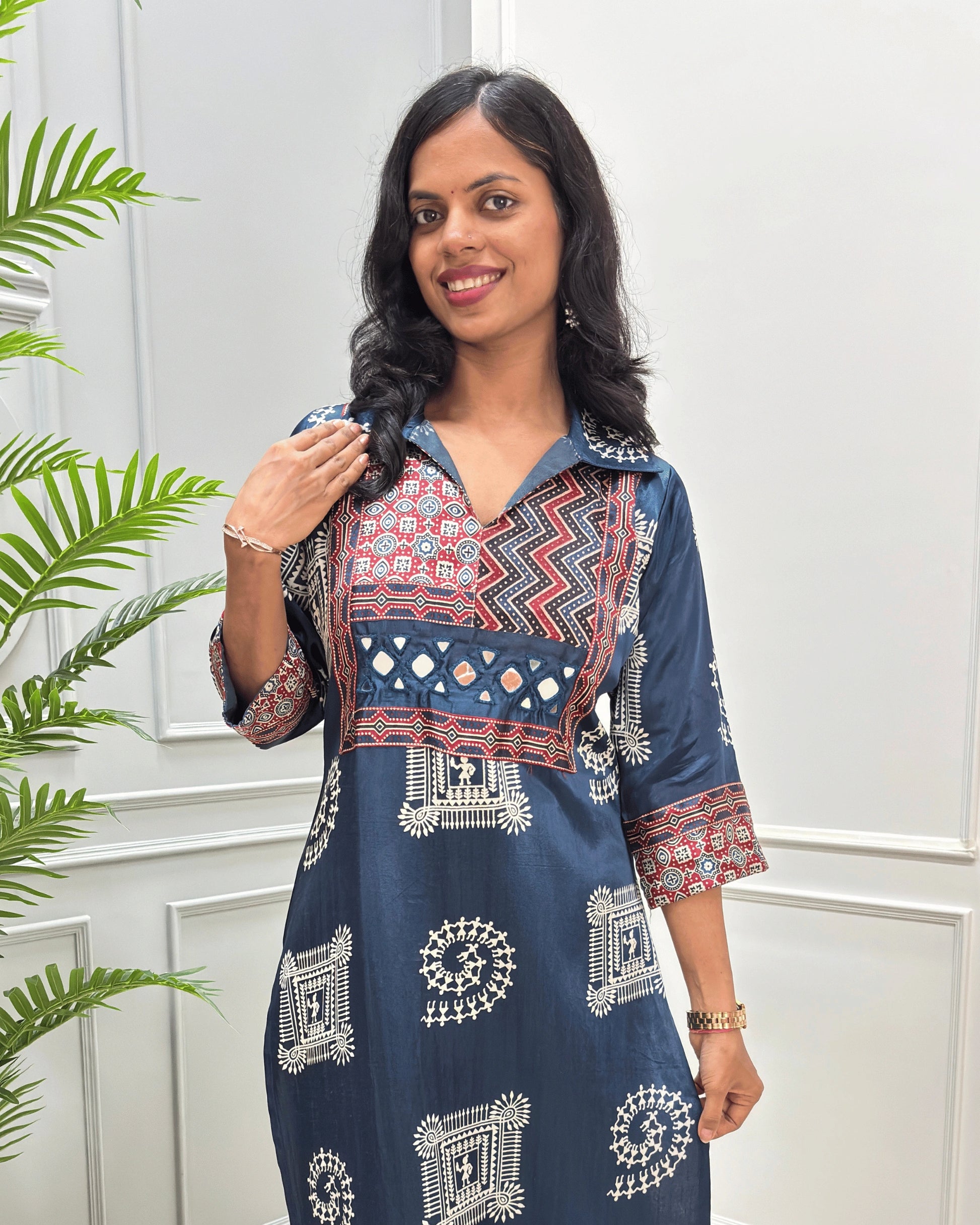 Close-up of Navy Blue Ajrakh Mashru silk Long kurti with multicolor geometric , warli print on front & sleeves along eith hand -mirror work on front , Collar V shaped neck patterns, standing in front of a white paneled wall with a green plant on the left.