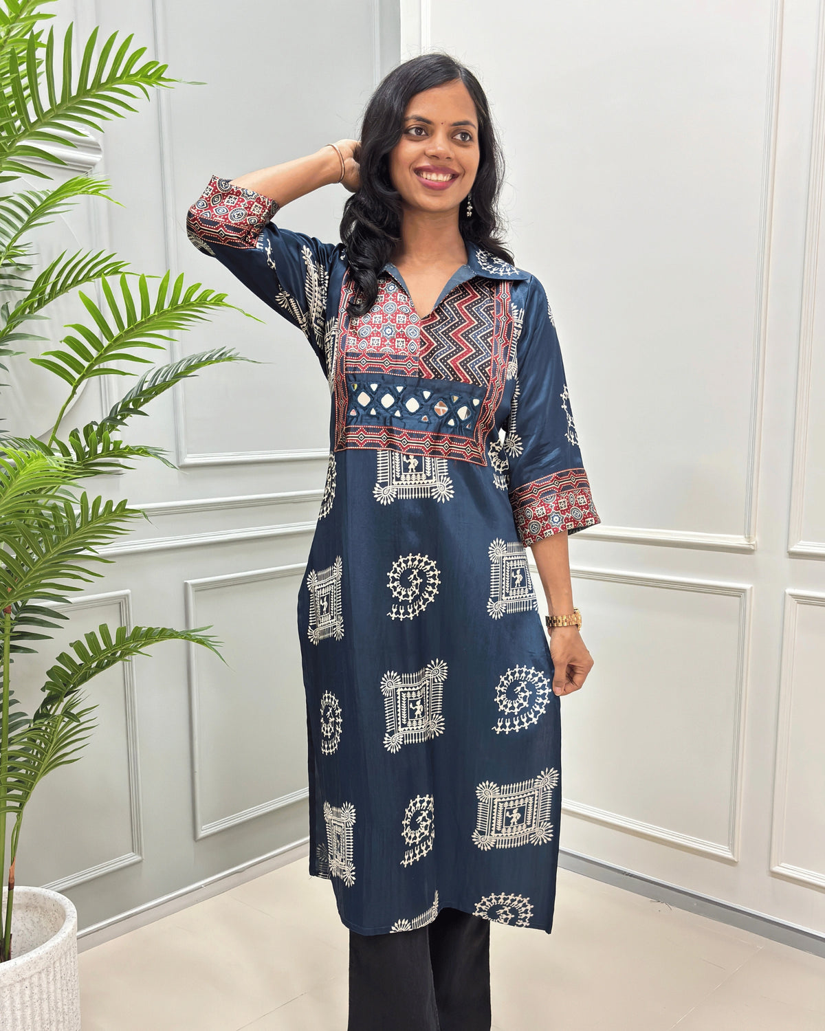 Front view of Navy blue Ajrakh pure Mashru Silk Long kurti with geometric print in red, white, and beige on Front & sleeves, standing in a bright room with a white paneled wall and a potted plant.