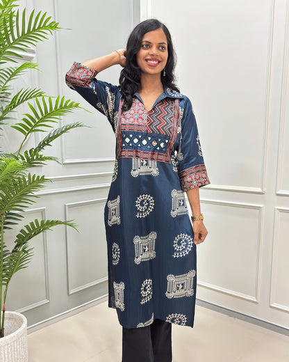 Front view of Navy blue Ajrakh pure Mashru Silk Long kurti with geometric print in red, white, and beige on Front & sleeves, standing in a bright room with a white paneled wall and a potted plant.