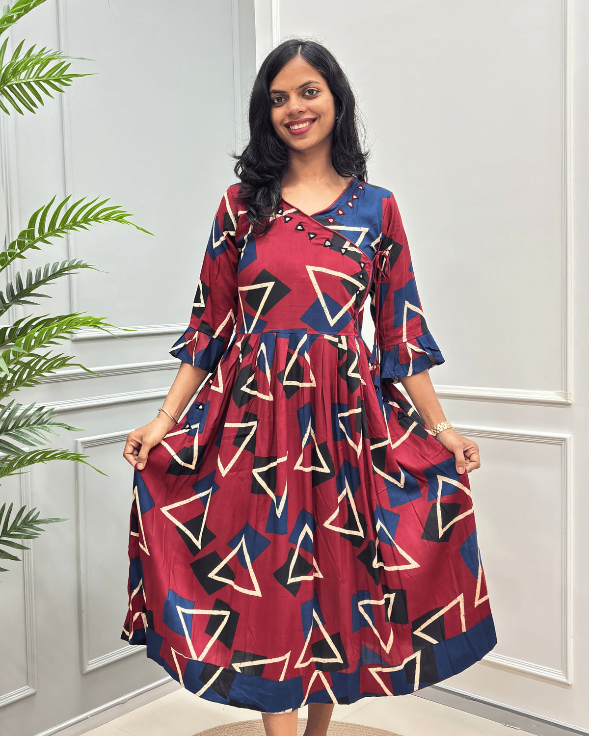 Front side of Red woman's cotton long-kurti with three-quarter sleeve, with a geometric triangle pattern in beige, navy, and black , standing in front of a white paneled wall with a green plant on the left.