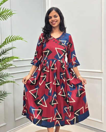 Front side of Red woman's cotton long-kurti with three-quarter sleeve, with a geometric triangle pattern in beige, navy, and black , standing in front of a white paneled wall with a green plant on the left.