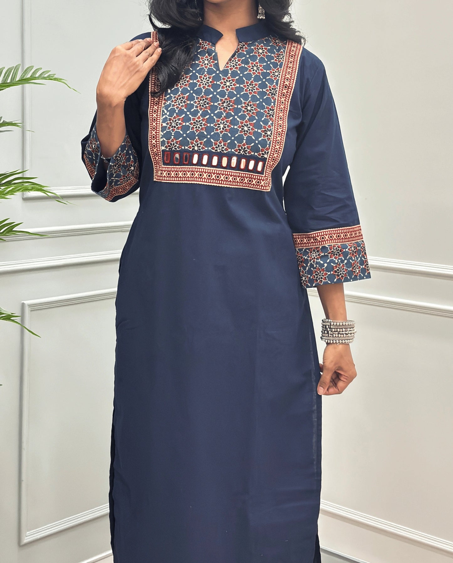 Close-up of Navy-Blue Hand-block Ajrakh Pure cotton Long kurti with three quarter sleeves with printed cuffs,  intricate black & beige geometric embroidery, with hand mirror work and thread work on border of mirror , with Mandarian collar V-neck and sleeve cuffs, standing against a light gray paneled wall.