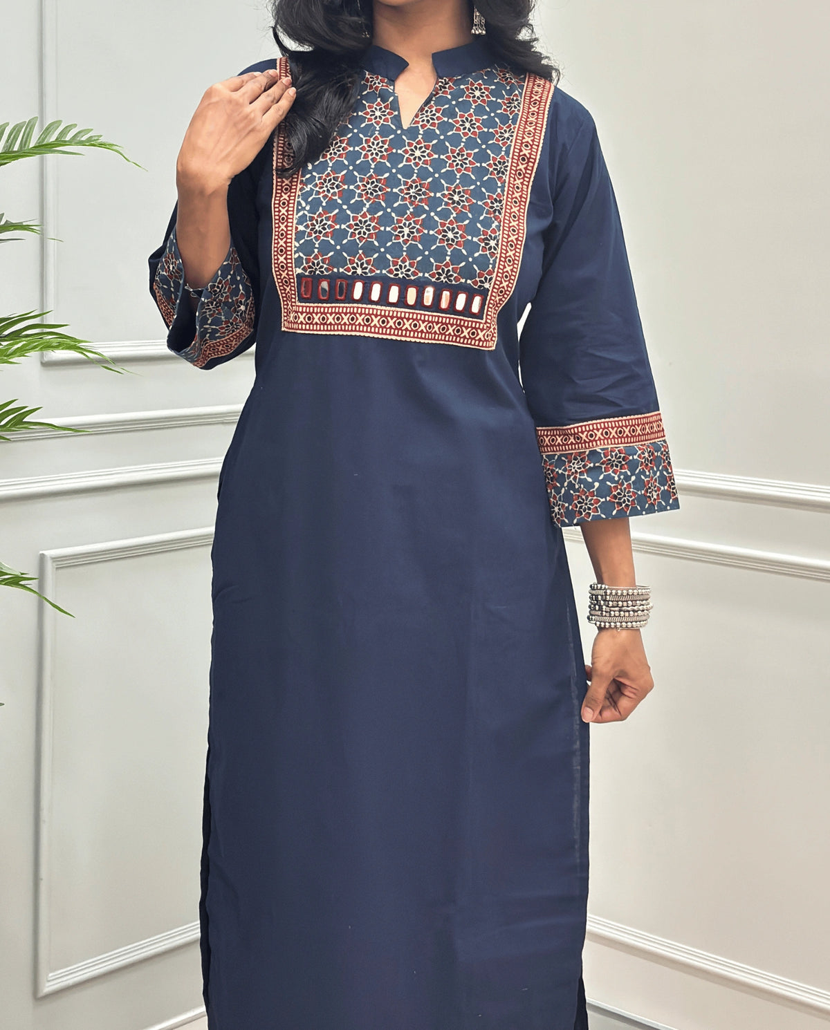 Close-up of Navy-Blue Hand-block Ajrakh Pure cotton Long kurti with three quarter sleeves with printed cuffs,  intricate black & beige geometric embroidery, with hand mirror work and thread work on border of mirror , with Mandarian collar V-neck and sleeve cuffs, standing against a light gray paneled wall.