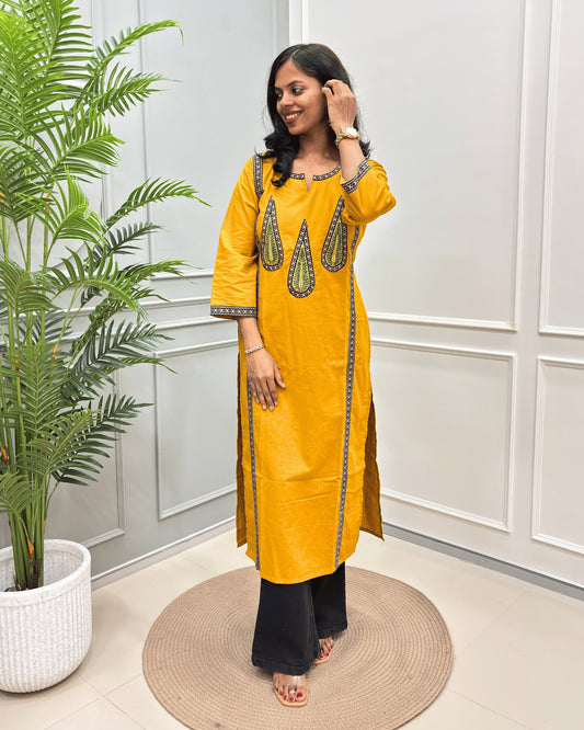 Yellow Women's Ajrakh Pure Cotton Long-kurti with Beige and Black Ajrakh print with green color  Palm-leaf detailing , paired with beige pants, standing in a room with a potted plant and paneled wall