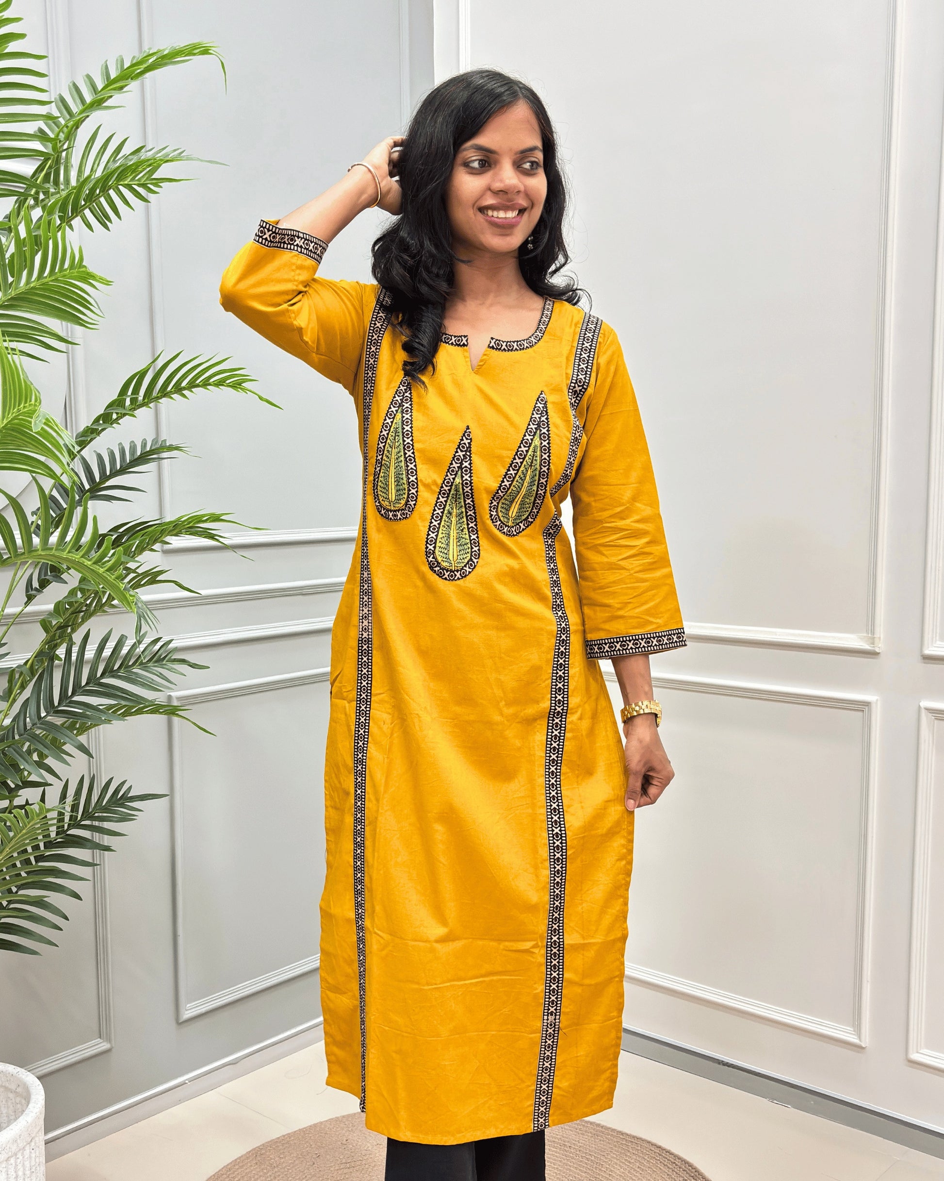 Front-view of Yellow Women's Hand-block Ajrakh Pure Cotton Long Kurti with beige and Black embroidered details on the V-neckline and borders, standing in a decorated room with a potted plant.