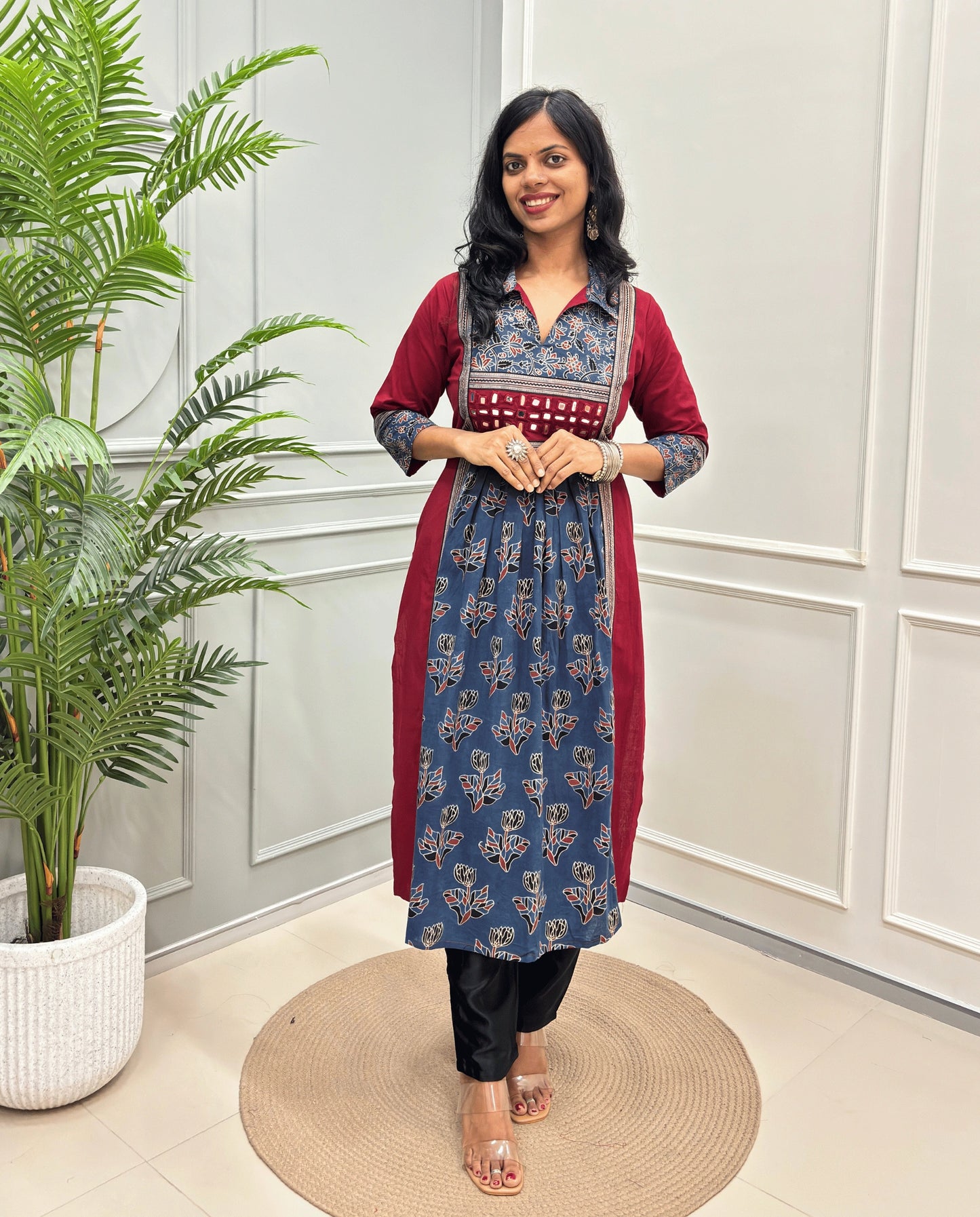 Blue Women Ajrakh Vegetable Dyed Plant Based Rayon Cotton Long Kurti Featured with Red & Blue Floral print , Aline pattern with side slight, paired with black pants, standing on a round woven mat in front of a white paneled wall with a potted plant on the left.