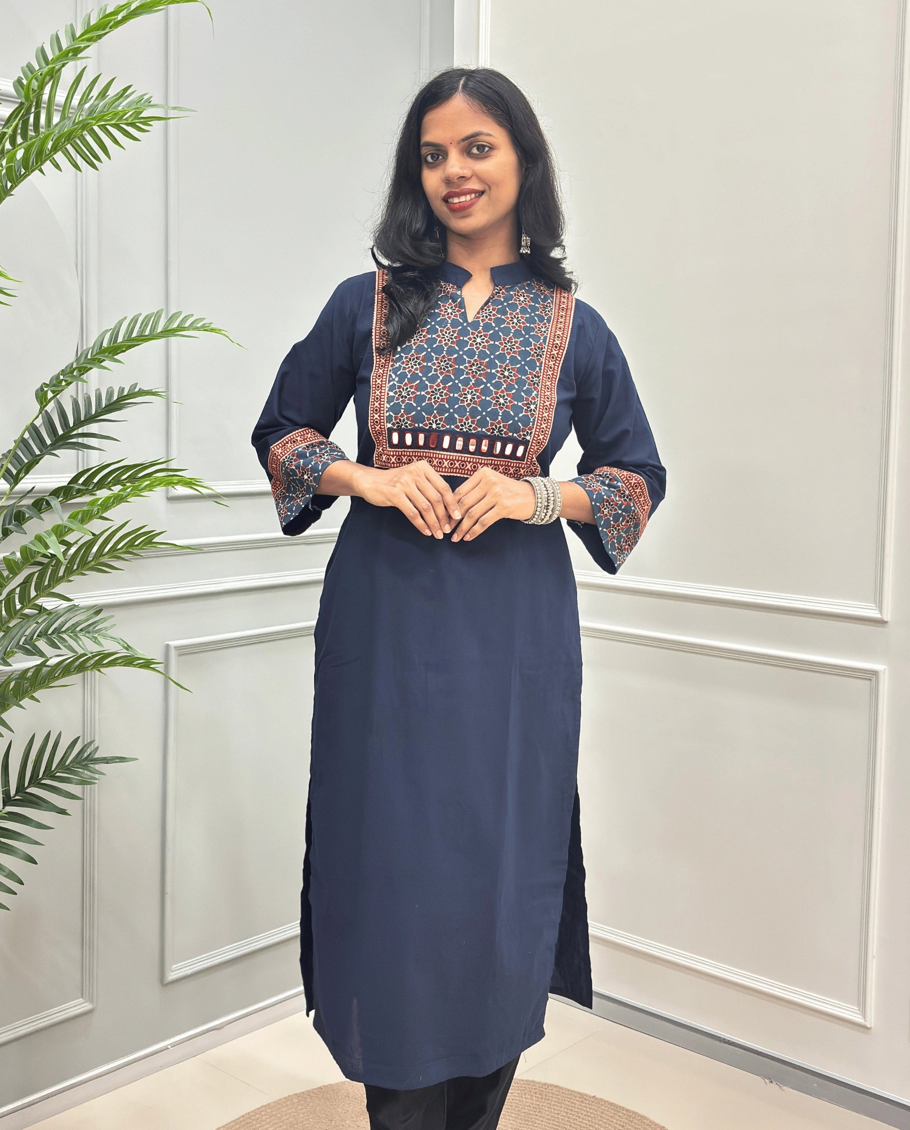 Front view of Navy-Blue Women's Hand-block Ajrakh Pure Cotton Long kurti with intricate green geometric print on the chest and sleeve cuffs, hand-mirror work on front , paired with black pants , standing on a round woven mat in front of a white paneled wall with a potted plant.