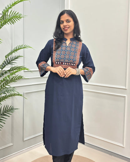 Front view of Navy-Blue Women's Hand-block Ajrakh Pure Cotton Long kurti with intricate green geometric print on the chest and sleeve cuffs, hand-mirror work on front , paired with black pants , standing on a round woven mat in front of a white paneled wall with a potted plant.