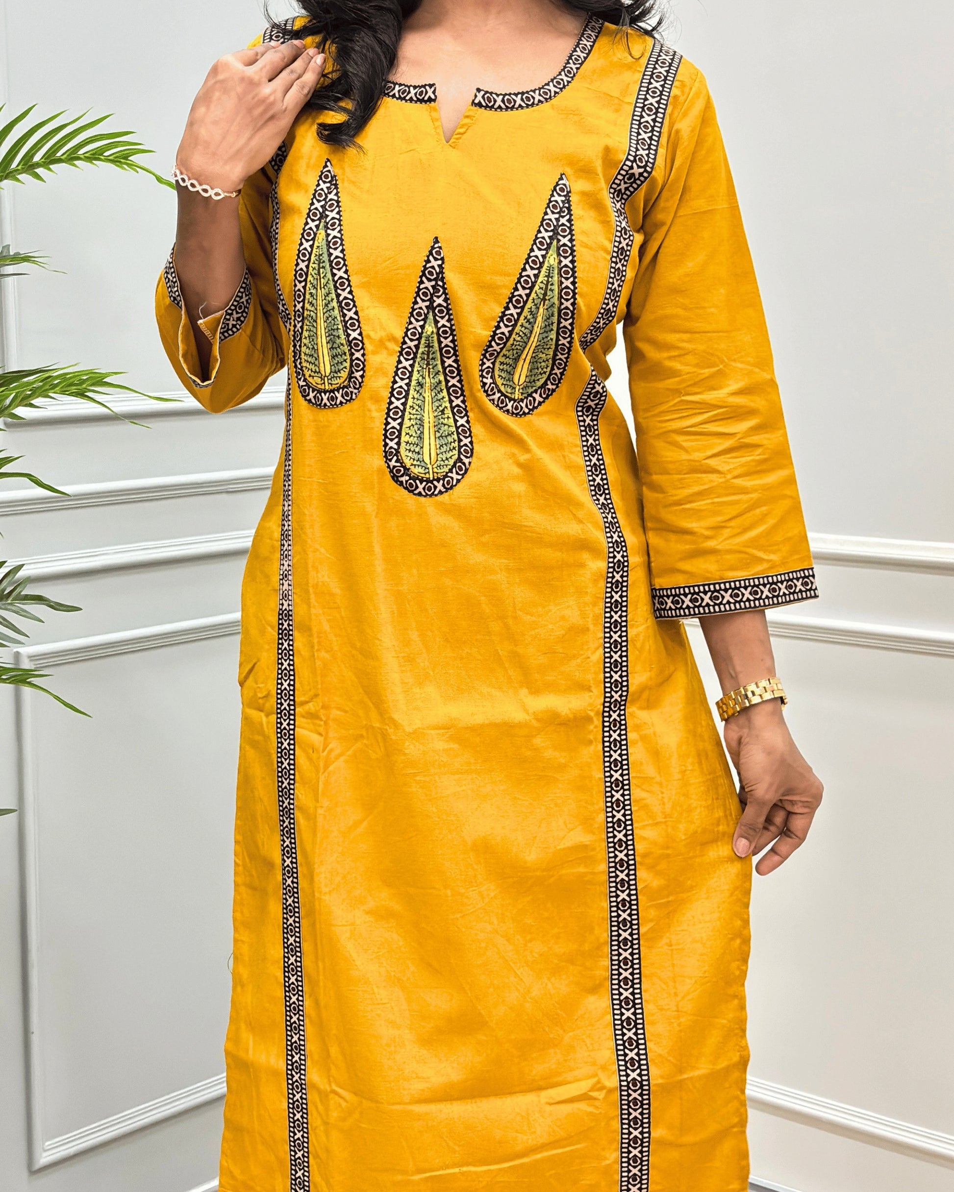 Close-up of Yellow Women's Hand-block Ajrakh Pure Cotton Long-kurti with three-quarter sleeve featured with intricate Green, Black and Yellow embroidered details on the neckline, cuffs, and front  panel, Along with Palm Leaf set against a light gray wall.