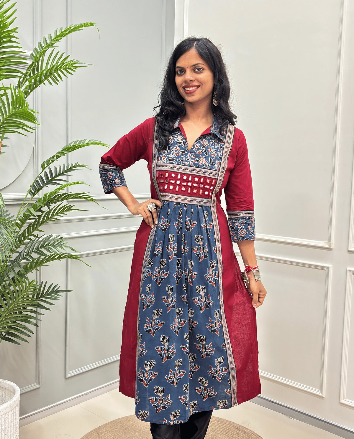 Front view of Blue women's Ajrakh plant based cotton Long-kurti featured with a red outer kurta and a blue printed inner kurta with floral motifs on front & sleeves , set against a white paneled wall with a green plant on the left.