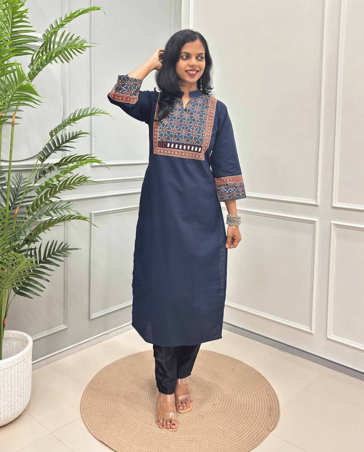 Navy-Blue Women's Hand-block Ajrakh Pure Cotton Long kurti with intricate white geometric print on the chest and sleeve cuffs, paired with black pants , standing on a round woven mat in front of a white paneled wall with a potted plant.