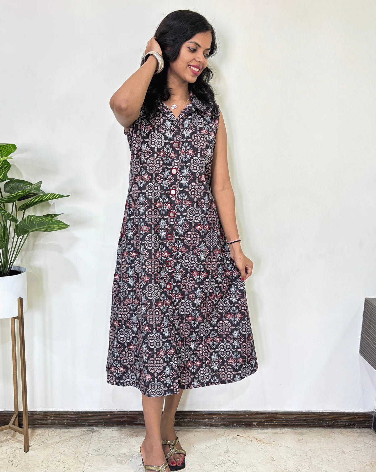 Black Women Ajrakh Vegetable dyed plant based cotton Long Kurti with a blue & red abstract print, Standing against a white wall with a plant to her left.