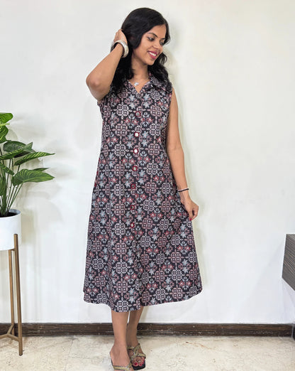 Black Women Ajrakh Vegetable dyed plant based cotton Long Kurti with a blue & red abstract print, Standing against a white wall with a plant to her left.