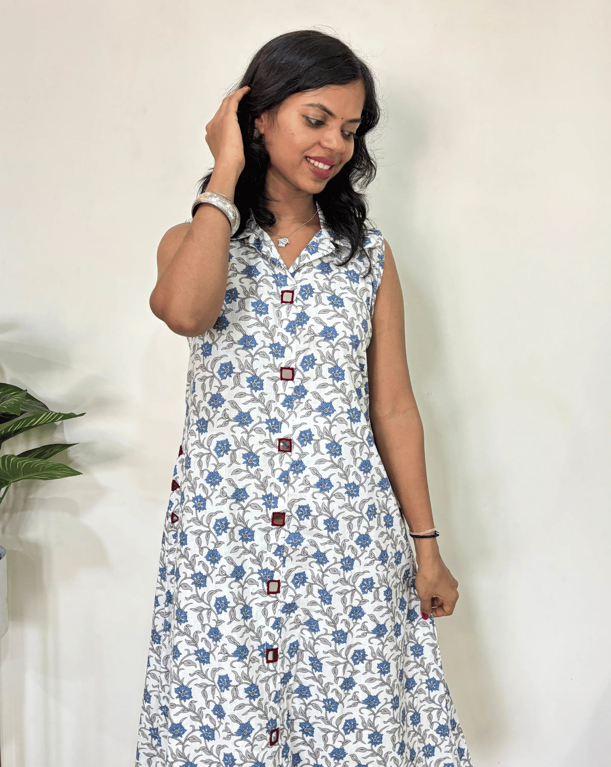 Close-up of White Women Ajrakh Vegetable dyed plant based cotton Long Kurti featured with Collar V Shaped neck with a blue abstract print, Hand-mirror work on Front & pockets, Standing against a white wall with a plant to her left.
