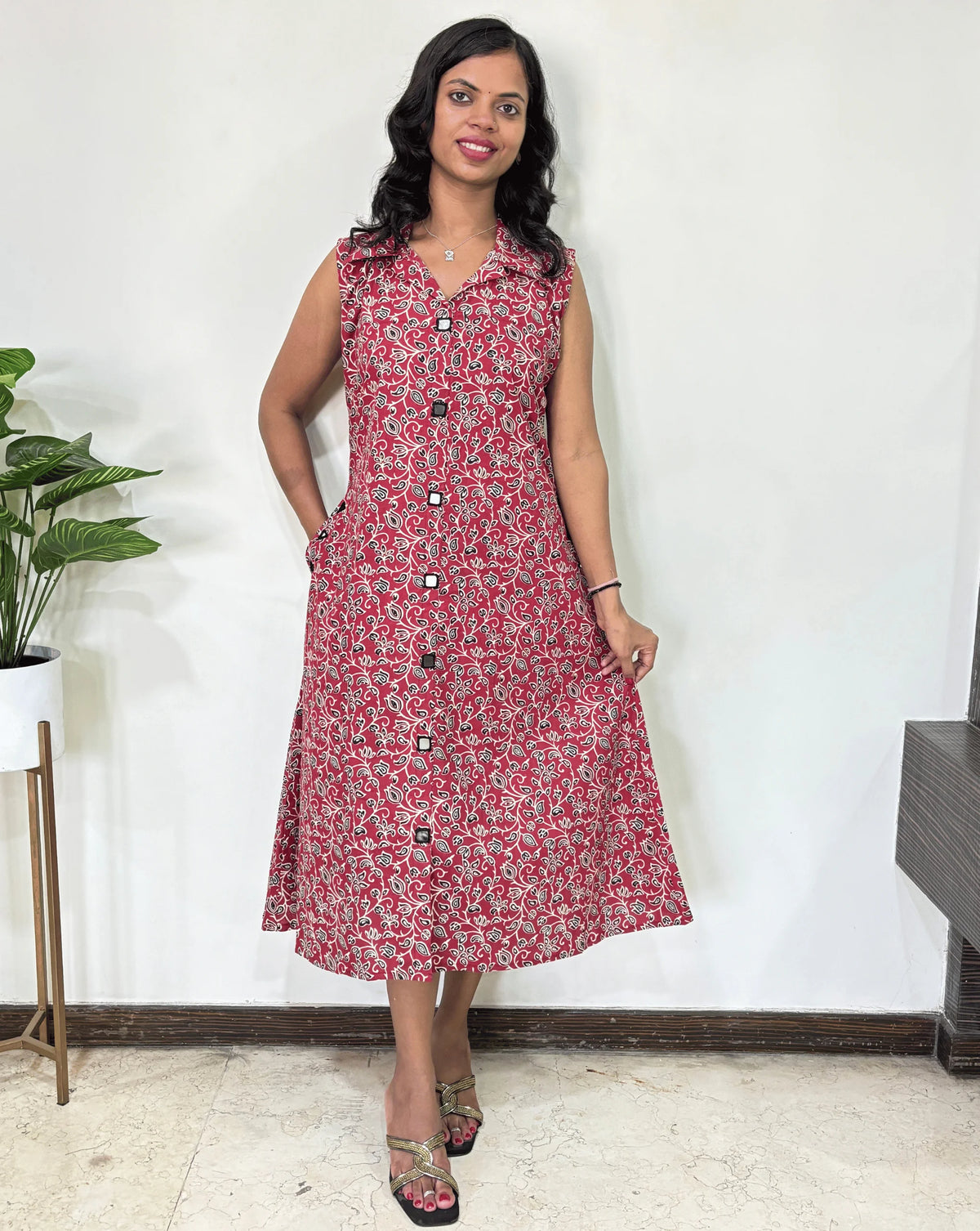 Red Women Ajrakh Vegetable dyed plant based cotton Long Kurti with a Black, & white abstract print, Standing against a white wall with a plant to her left.