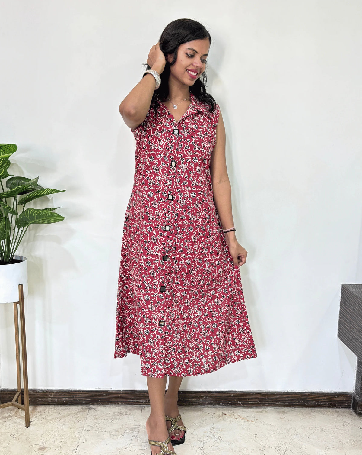 Close-up of Red Women Ajrakh Vegetable dyed plant based cotton Long Kurti Featured Collar V-shaped neck with a Black, & white abstract print, Hand-mirror work on front & pockets,  Standing against a white wall with a plant to her left.