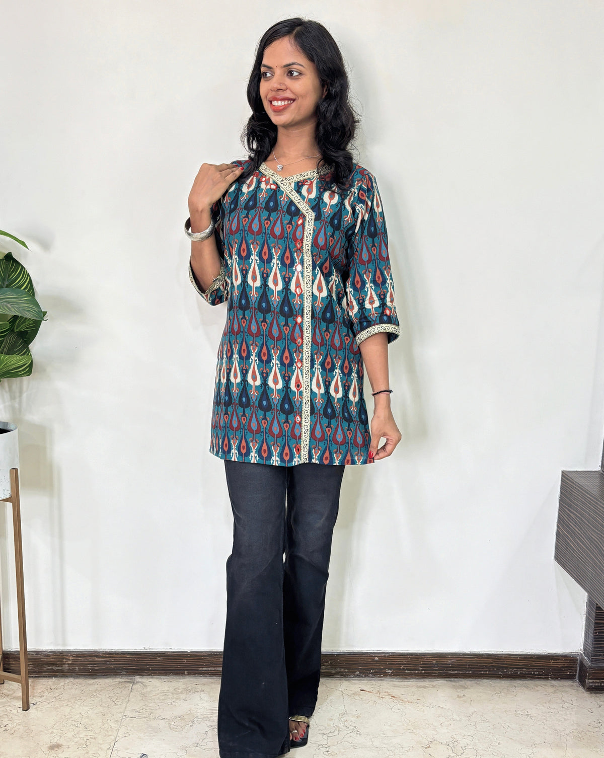 Green Women's Ajrakh vegetable dyed plant based cotton Short kurti with a multi-colour patola print with white border in Y shape on front & sleeves cuffs, paired with Black flared jeans, Standing in front of white wall.