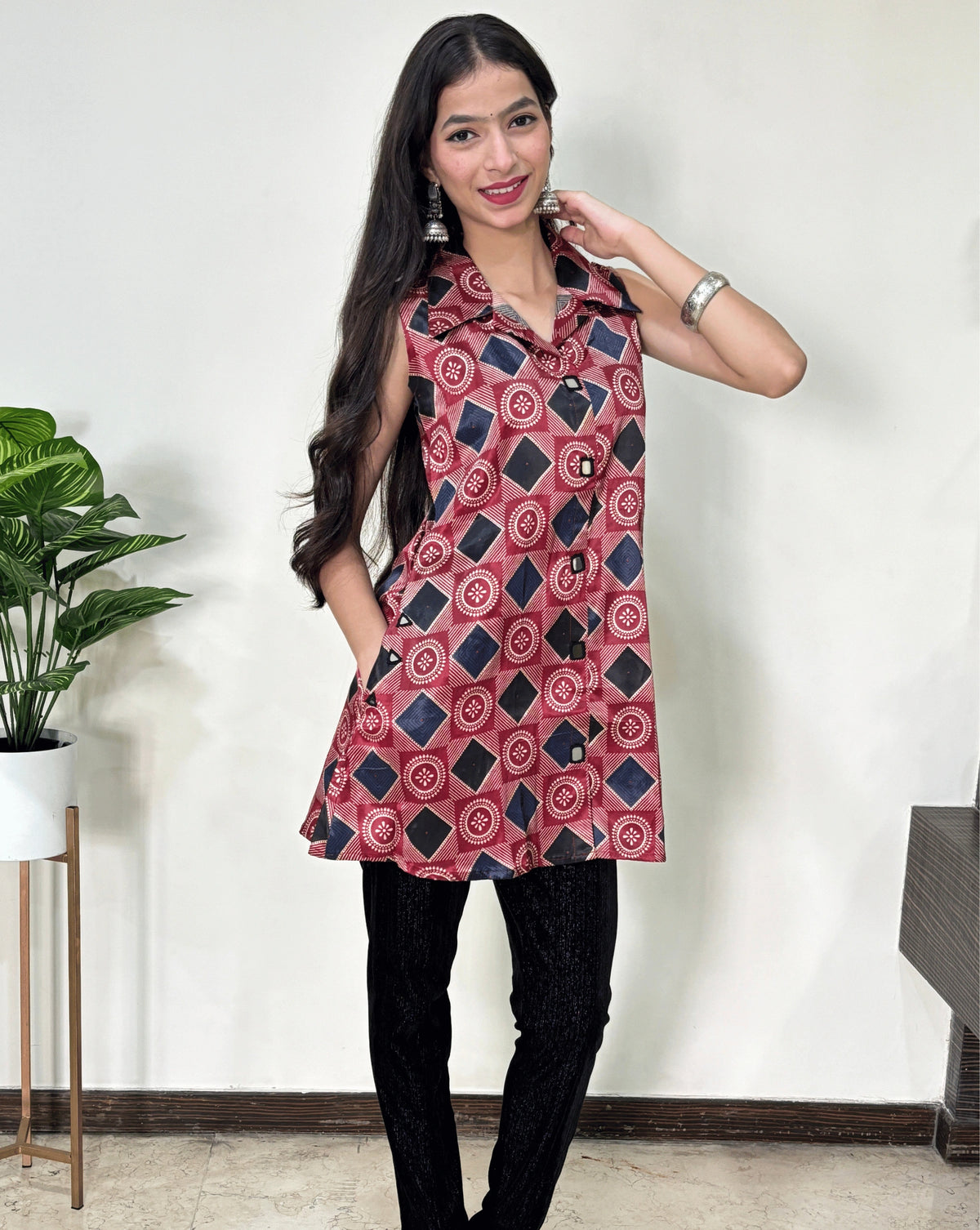 Red Women's Ajrakh pure Mashru Silk Short Kurti with a Bandhani Geometric print Abstract is blue & white, & A-line pattern, paired with black pant, Standing in front of white wall, on left side Plant pot.
