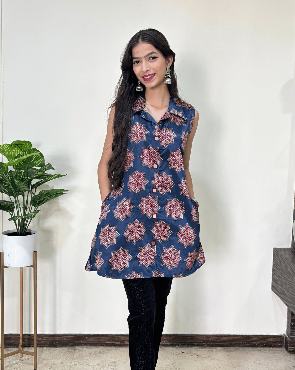 Close-up of Blue Women's Ajrakh pure Mashru Silk Short Kurti featured with collar neck & sleeveless, with a Blue base & an abstract is Red Star print & A-line pattern,2 side pockets, Hand-mirror work on front look-a-like buttons, paired with black pant, Standing in front of white wall, on left side Plant pot.