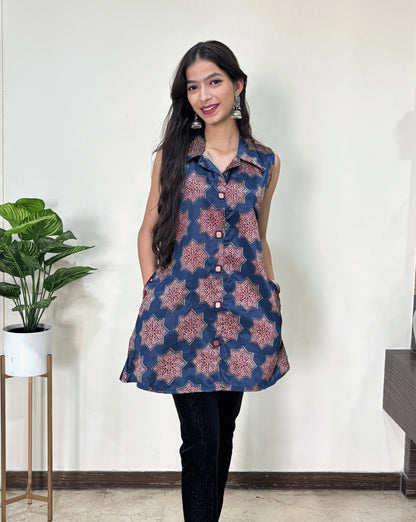 Close-up of Blue Women's Ajrakh pure Mashru Silk Short Kurti featured with collar neck & sleeveless, with a Blue base & an abstract is Red Star print & A-line pattern,2 side pockets, Hand-mirror work on front look-a-like buttons, paired with black pant, Standing in front of white wall, on left side Plant pot.