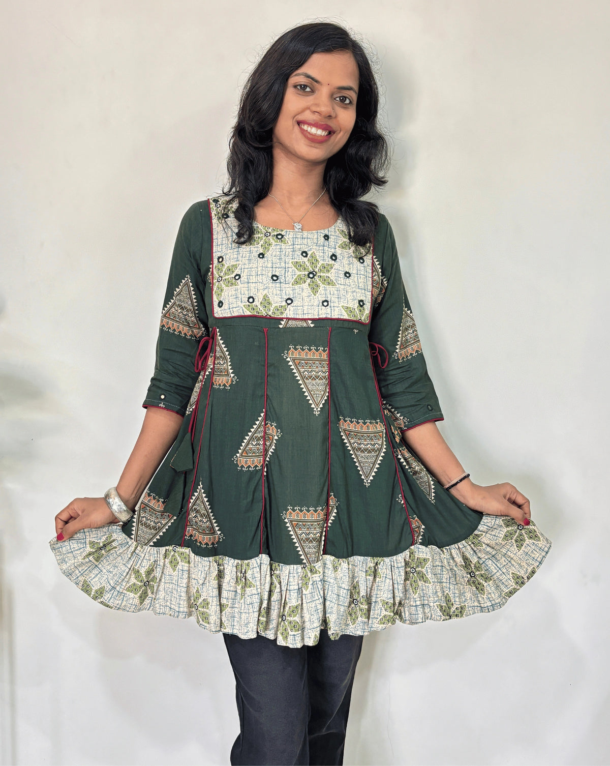 Close-up of Green Ajrakh plant based cotton Short kurti with geometric patterns, a Beige brush print yoke & bottom, Red panel strip with lace on side, Hand-mirror work on round neck paired with dark pants against a plain light background.