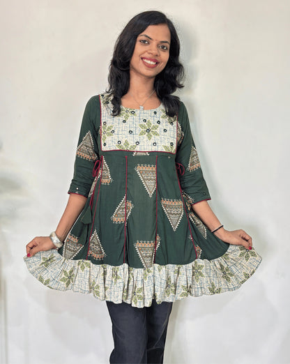 Close-up of Green Ajrakh plant based cotton Short kurti with geometric patterns, a Beige brush print yoke & bottom, Red panel strip with lace on side, Hand-mirror work on round neck paired with dark pants against a plain light background.