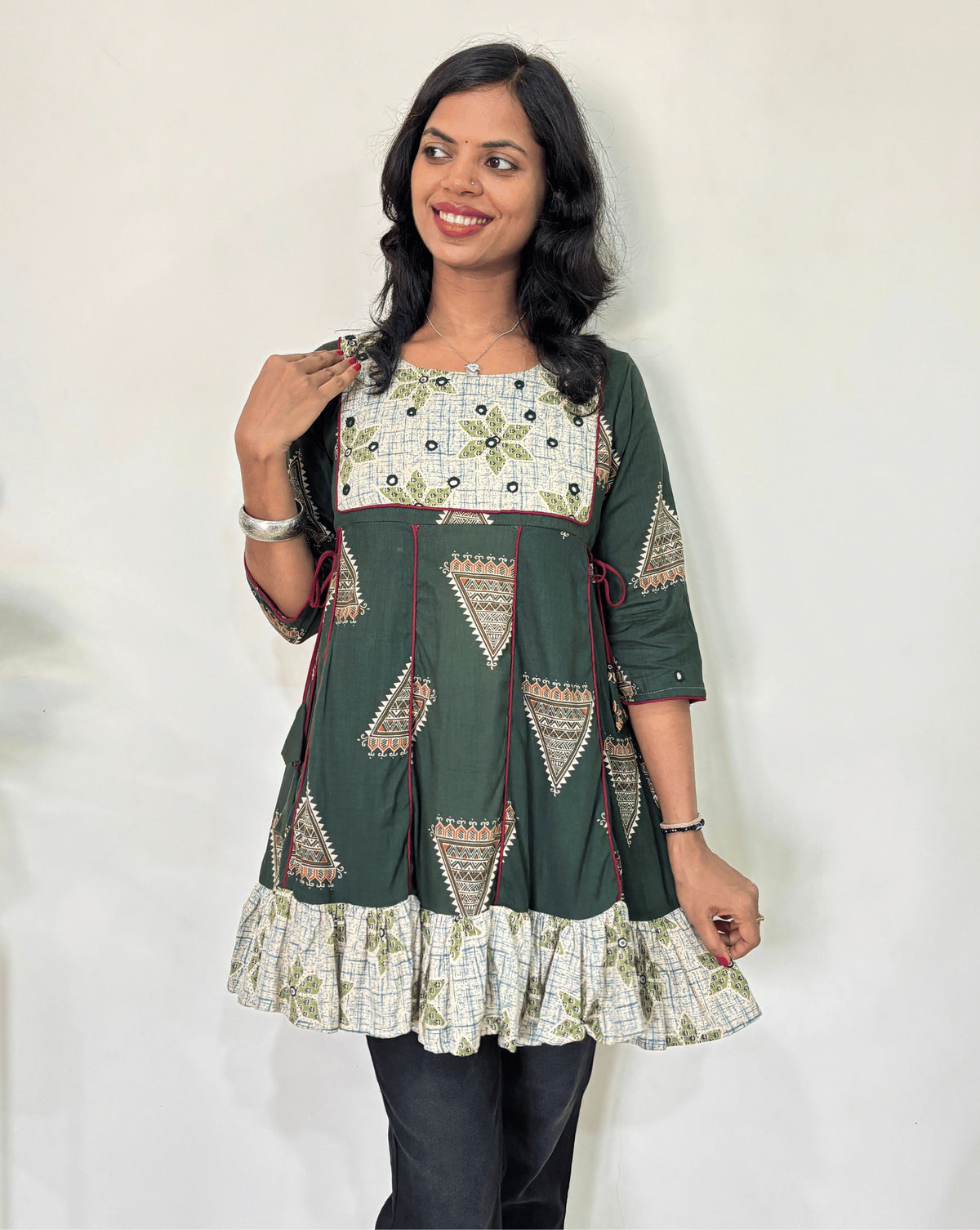 Front veiw of Green Ajrakh Plant based cotton Short kurti featured with Green and grey geometric print & aline frill patterned dress,  standing against a white wall.