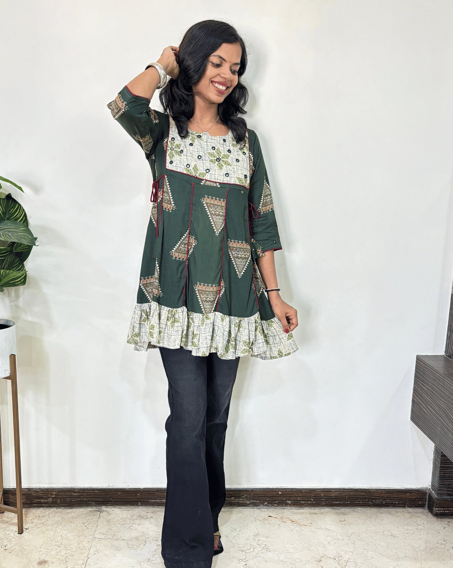 Green Women's Ajrakh Vegetable Dyed Plant based Cotton Short Kurti with three quarter sleeves . White & Green base & geometric print on Sleeves & front , Standing in front of white wall. 