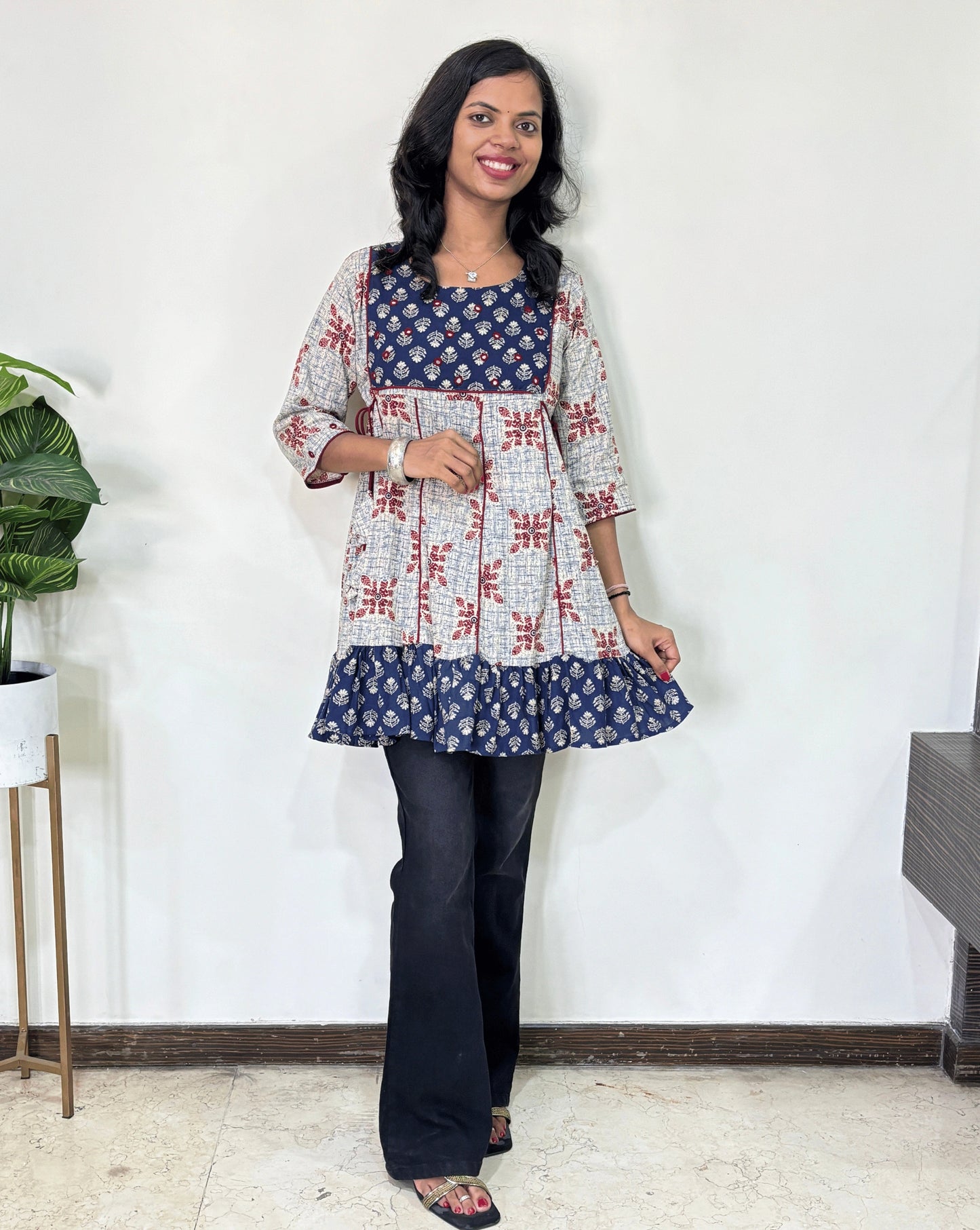 White Women's Ajrakh Vegetable Dyed Plant based Cotton Short Kurti with three quarter sleeves . Blue & White base & geometric print on Sleeves & front , Standing in front of white wall. 