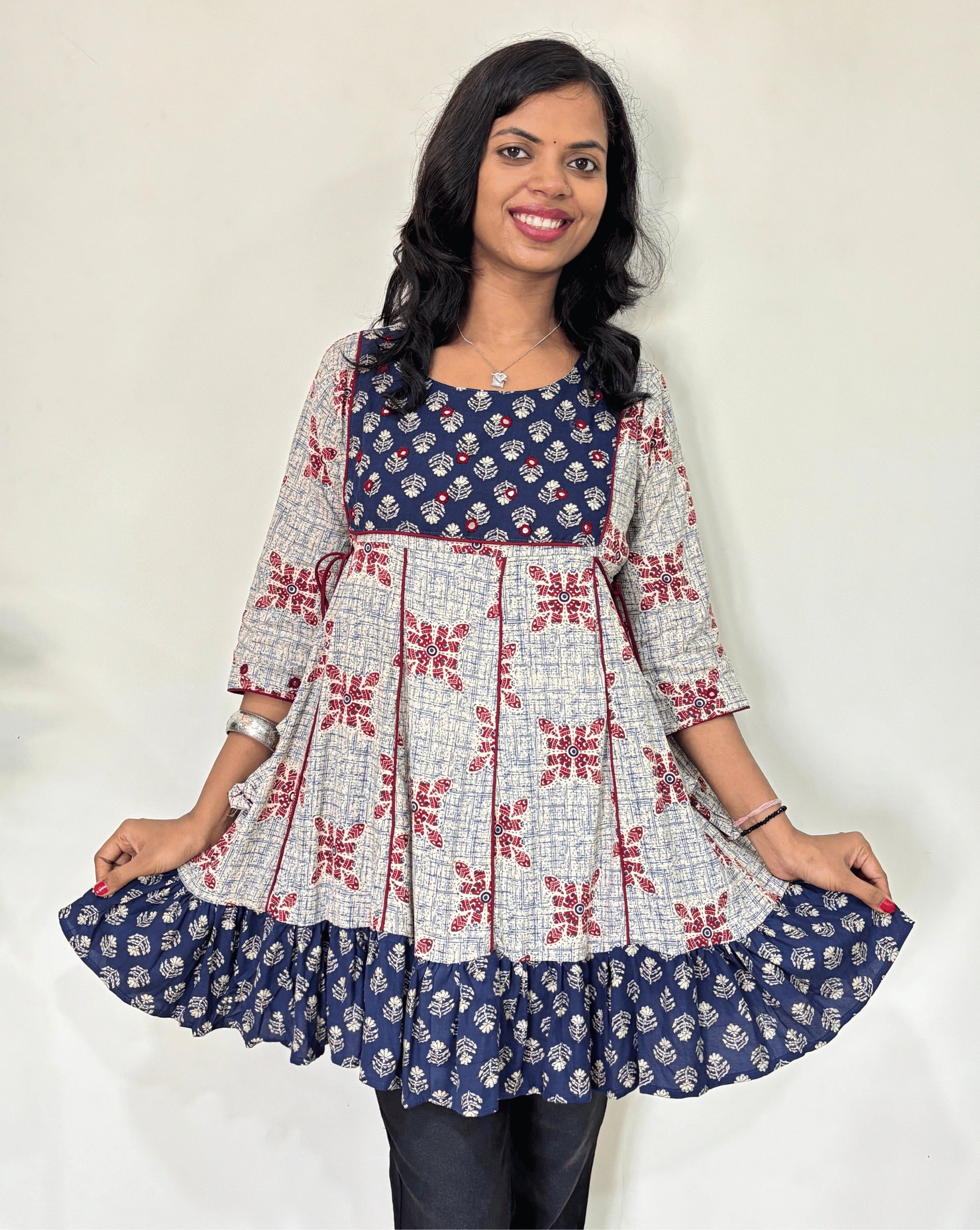 Close-up of white Ajrakh plant based cotton Short kurti with geometric patterns, a blue brush print yoke & bottom, Red panel strip with lace on side, Hand-mirror work on round neck paired with dark pants against a plain light background.