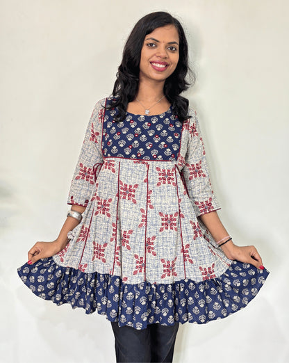 Close-up of white Ajrakh plant based cotton Short kurti with geometric patterns, a blue brush print yoke & bottom, Red panel strip with lace on side, Hand-mirror work on round neck paired with dark pants against a plain light background.