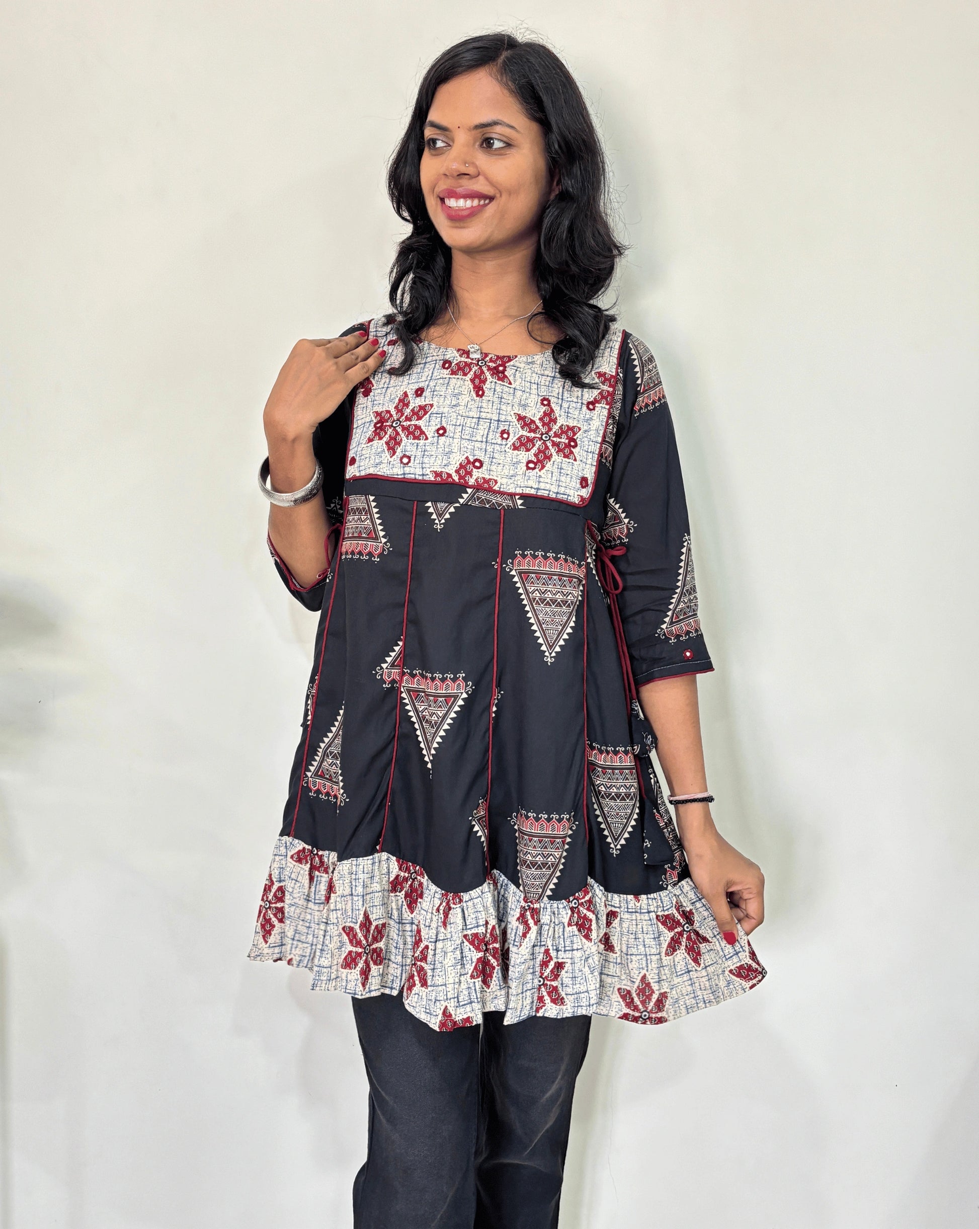 Close-up of Black Ajrakh plant based cotton Short kurti with geometric patterns, a Grey brush print yoke & bottom, Red panel strip with lace on side, Hand-mirror work on round neck paired with dark pants against a plain light background.