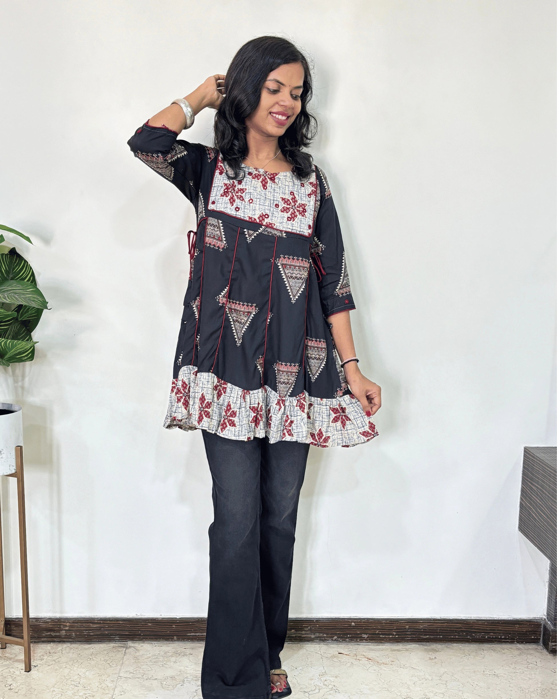 Front veiw of Black Ajrakh Plant based cotton Short kurti featured with Grey and Black geometric print & aline frill patterned dress,  standing against a white wall. 