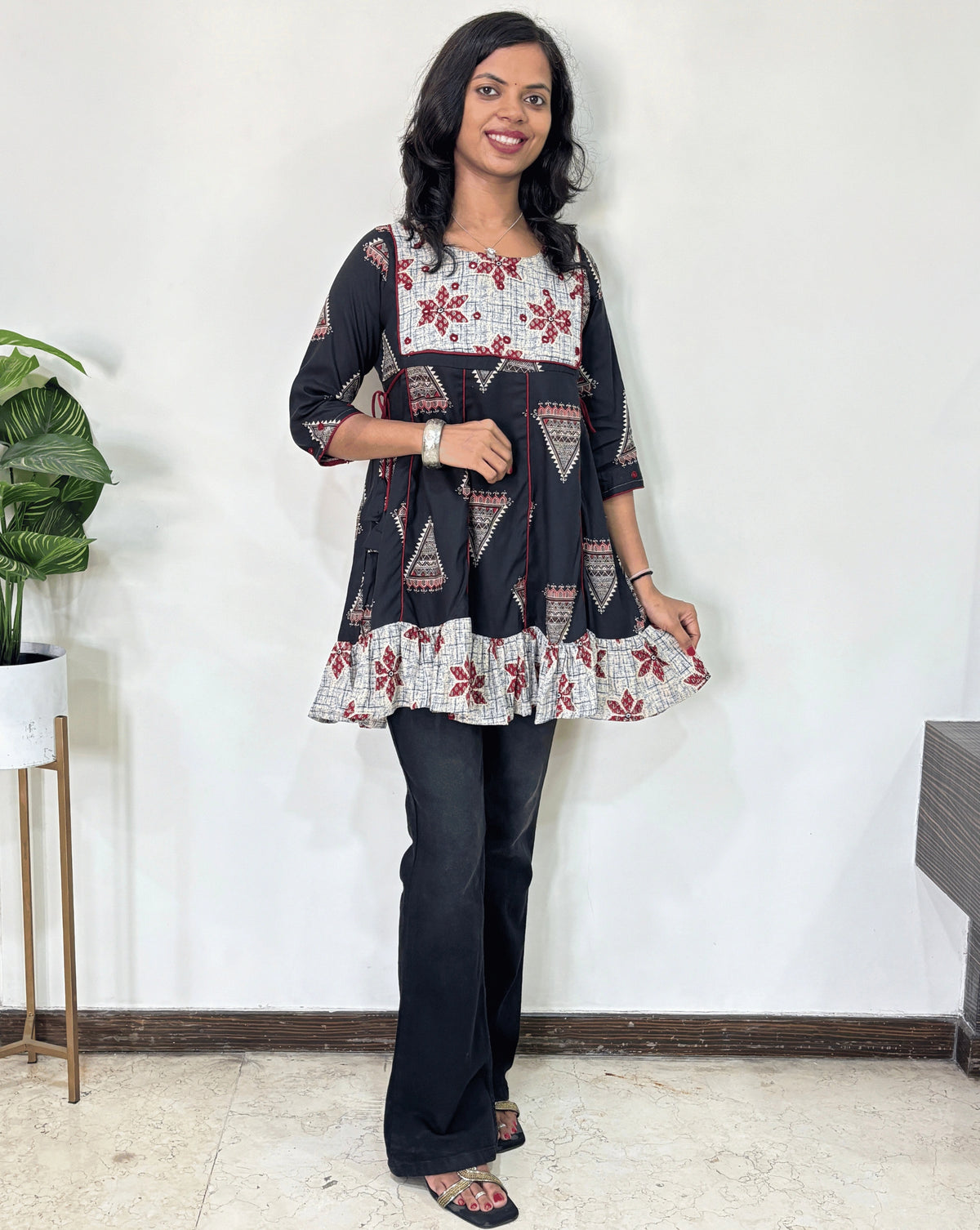 Black Women's Ajrakh Vegetable Dyed Plant based Cotton Short Kurti with three quarter sleeves . Grey & Black base & geometric print on Sleeves & front , Standing in front of white wall. 